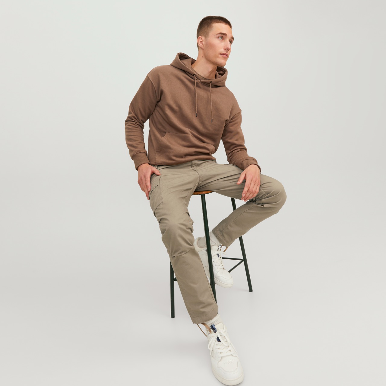Homem sentado com hoodie castanho, calças bege e ténis brancos em fundo branco