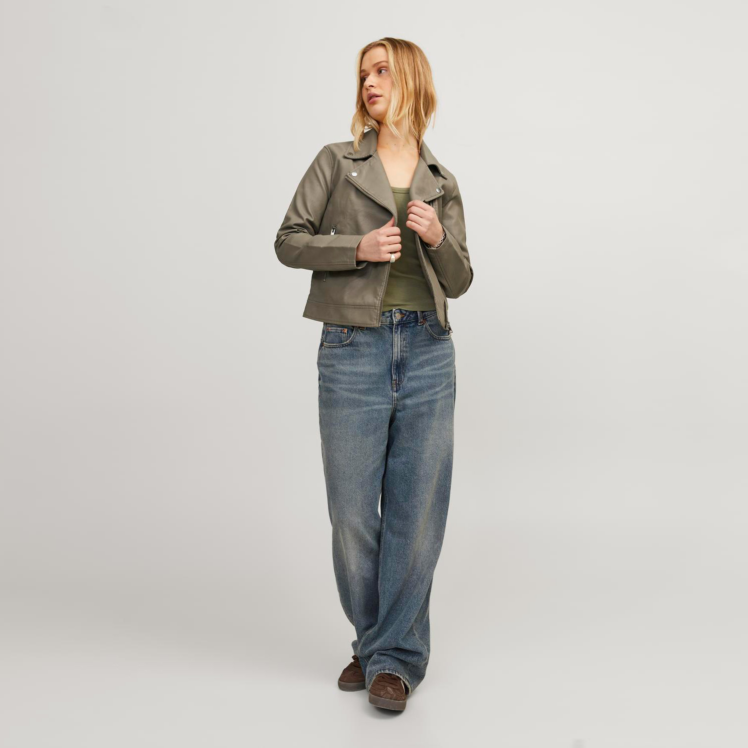 mulher com casaco cinza, camisa verde, jeans largos azuis e sapatos castanhos, fundo branco