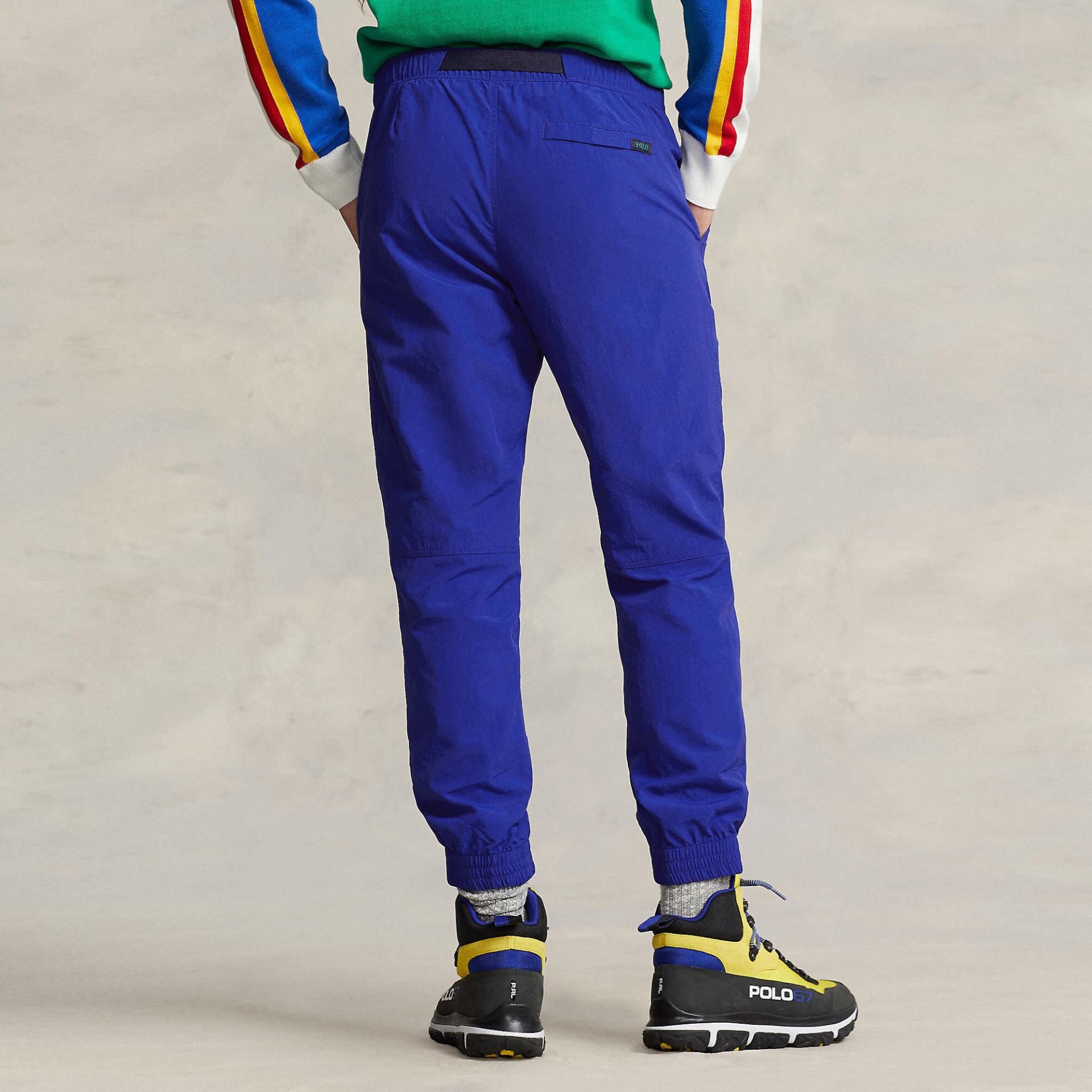 Calças desportivas azuis com bainha elástica e bolso traseiro com etiqueta POLO, usadas com camisola colorida e botas POLO67.