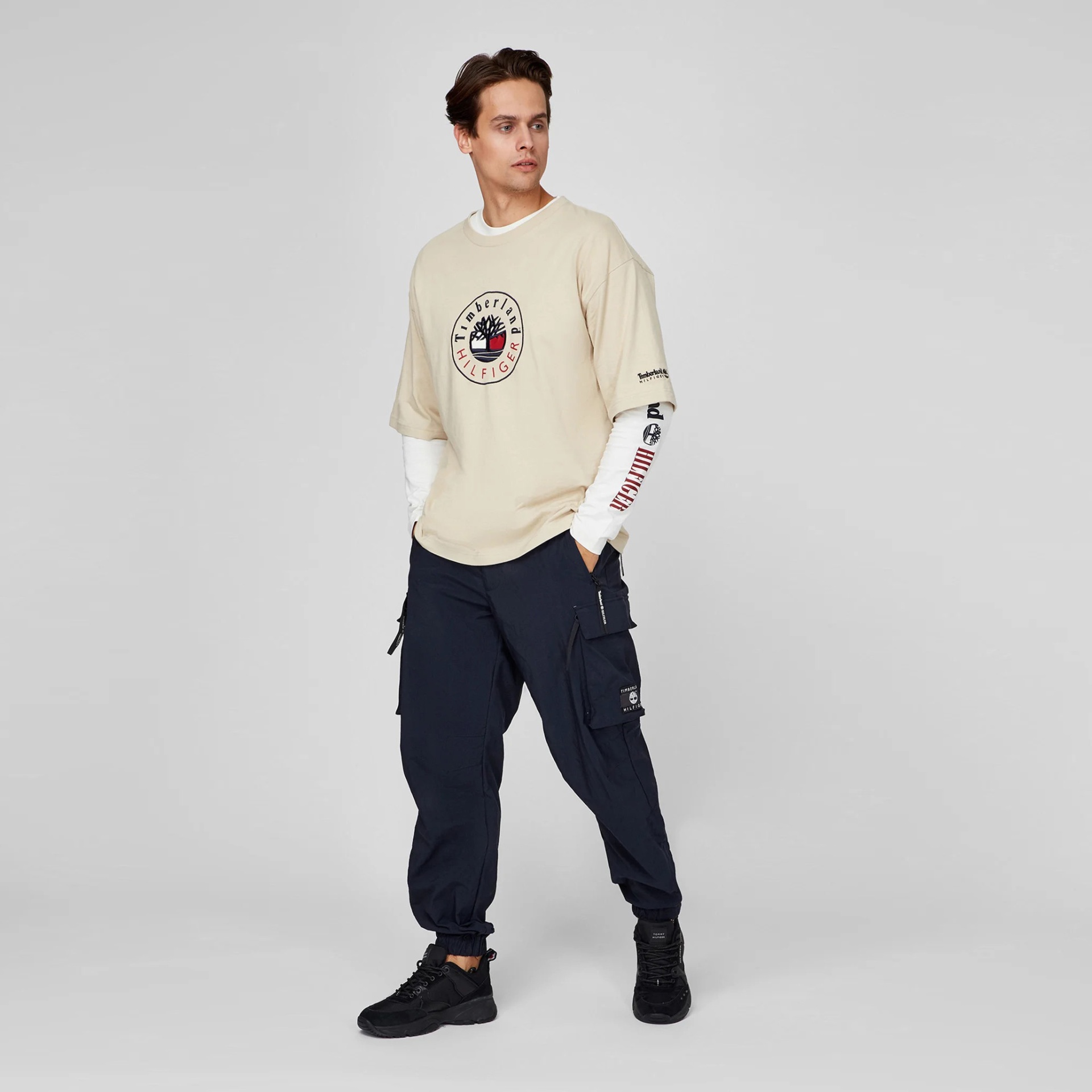 Homem com t-shirt bege, camisa branca de manga comprida, calças cargo azuis escuras e ténis pretos em fundo cinzento