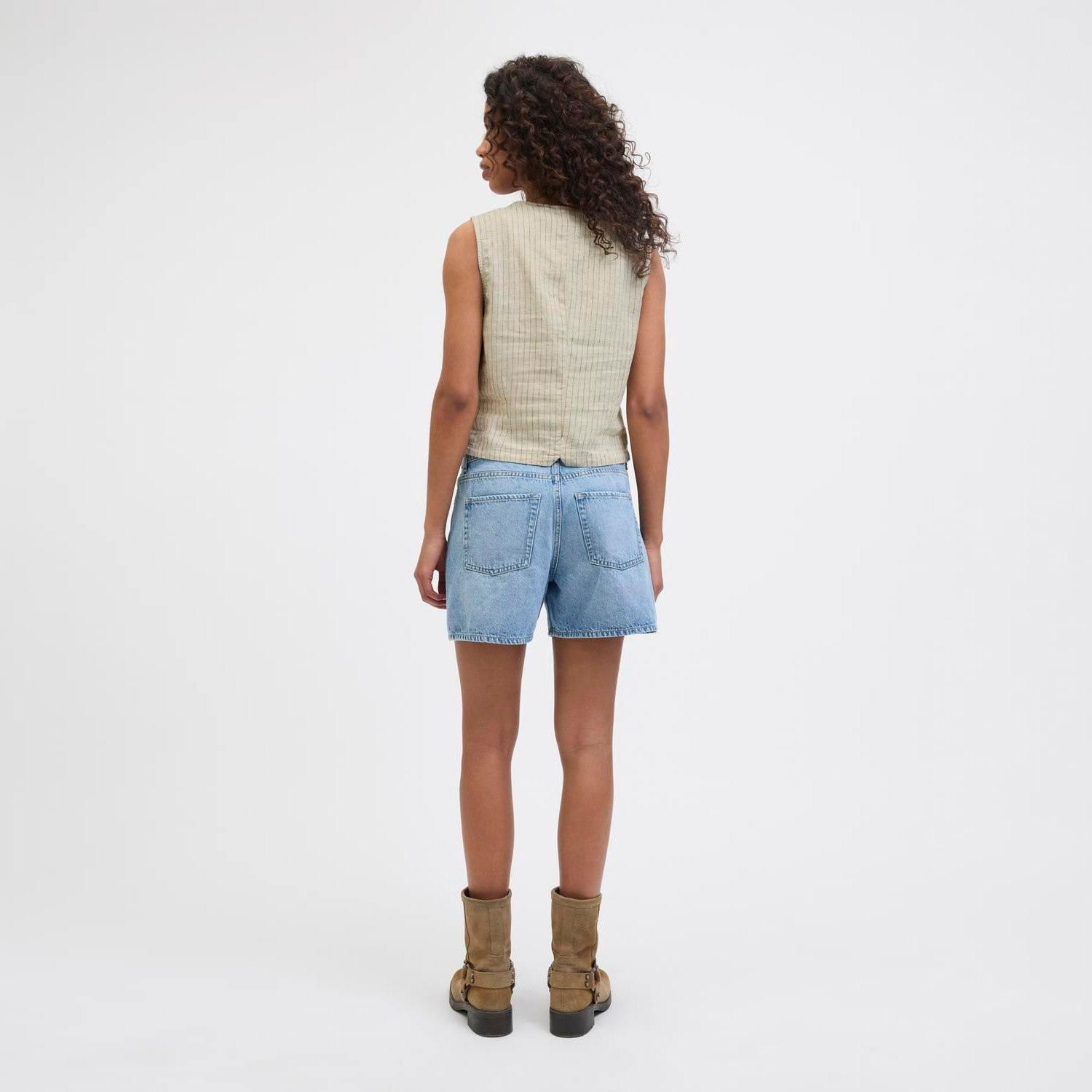 Mulher de costas vestindo colete bege, calções de ganga e botas castanhas