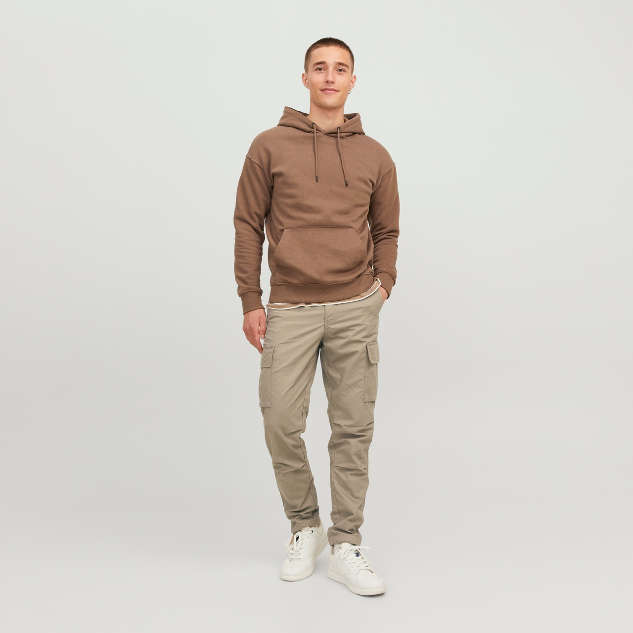 Homem com hoodie castanho, calças cargo bege e sapatilhas brancas em fundo branco