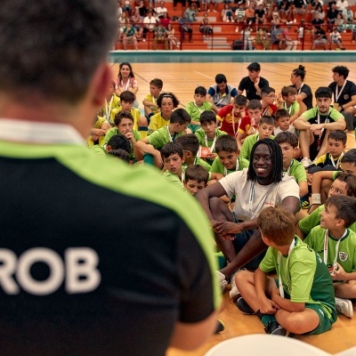 A nossa paixão pelo desporto ganha uma nova dimensão: Grupo Recreativo Olival Basto (GROB) Parceiro época 23/24 Camisola preta e verde com GROB nas costas em ginásio com crianças em uniformes verdes e amarelos sentadas no chão