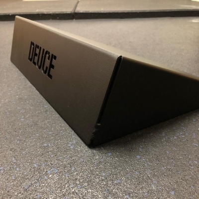 Suporte triangular marrom escuro com palavra DEUCE em preto sobre chão cinza.