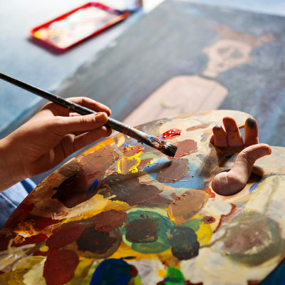 Paleta colorida de pintura com mãos segurando pincel e tela pintada ao fundo