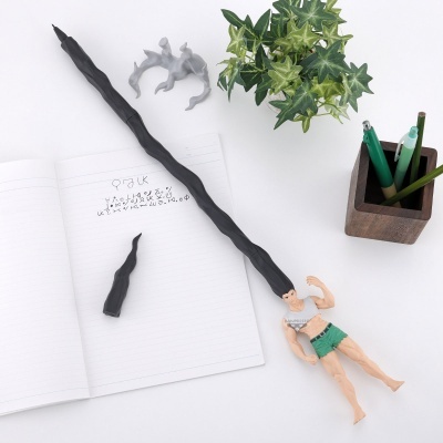 Figura de ação masculina com cabelo preto comprido sobre mesa com caderno, planta, canetas e acessório cinzento