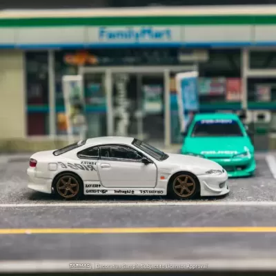 Modelo de carro desportivo branco com rodas douradas e adesivos estacionado frente a loja FamilyMart com carro verde ao fundo