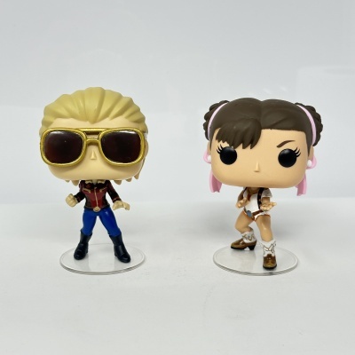 Duas figuras Funko Pop em fundo branco, uma loira com óculos e outra sorridente com tranças.