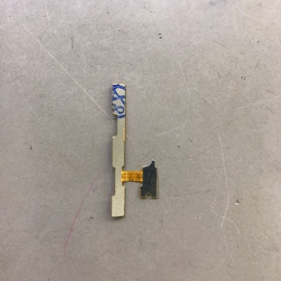 Componente eletrónico pequeno com fita flexível amarela e partes metálicas cinzentas sobre superfície riscada