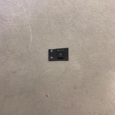 Componente eletrónico preto quadrado com lente central sobre superfície cinzenta