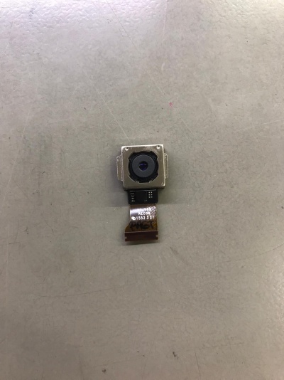 Pequena câmara de telemóvel com lente preta em estrutura metálica quadrada e fita flexível com texto visível numa superfície cinzenta.