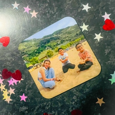 Íman com fotografia de três crianças na praia rodeado por confetes coloridos em fundo escuro.