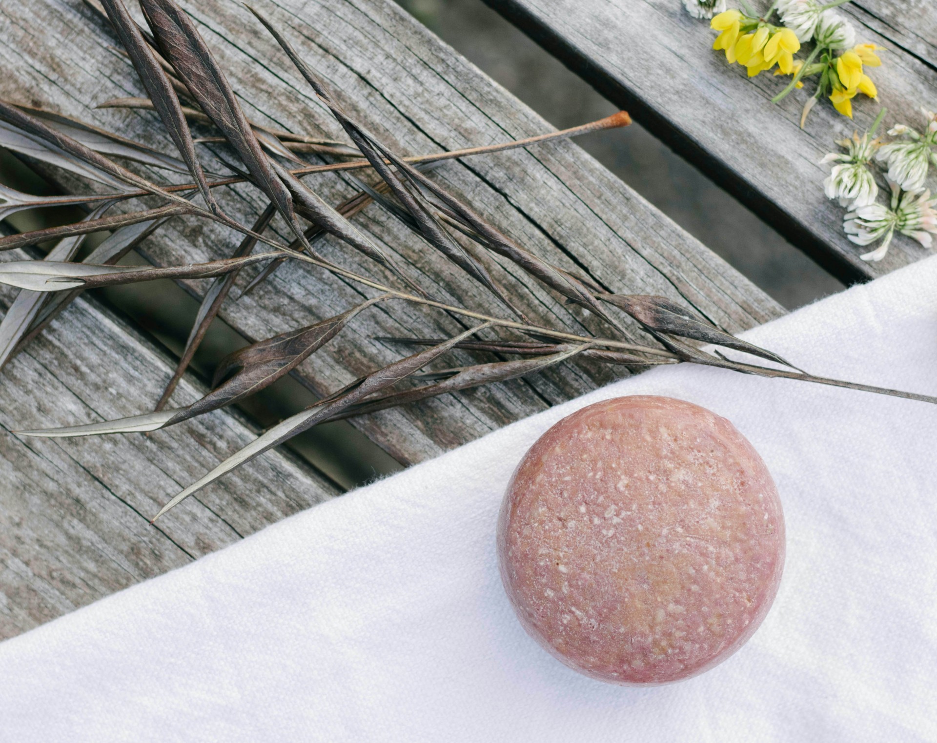 Barra redonda de xampu sólido rosa sobre pano branco e madeira com ramos secos e flores