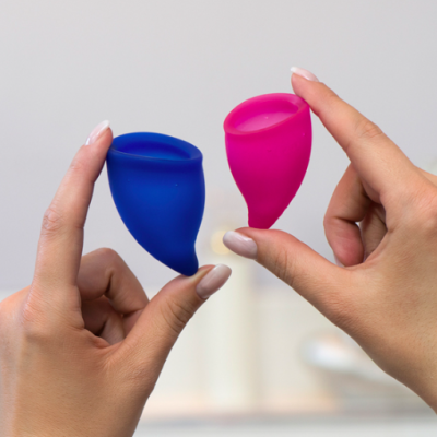 Dois copos menstruais azul e rosa segurados por mãos femininas