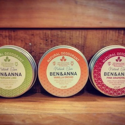 Três desodorizantes naturais Ben & Anna em latas coloridas sobre madeira.