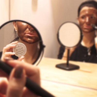 Mulher a aplicar máscara facial com recipiente branco refletida em espelho de mesa