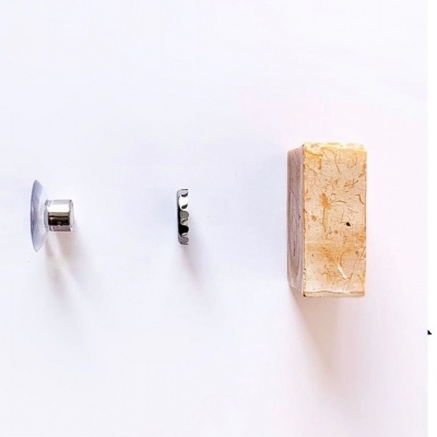 Três objetos alinhados: suporte metálico com ventosa, peça metálica pequena ondulada, bloco retangular creme com textura.