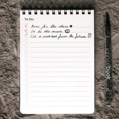 Bloco de notas com lista escrita à mão e caneta preta numa superfície de areia cinzenta