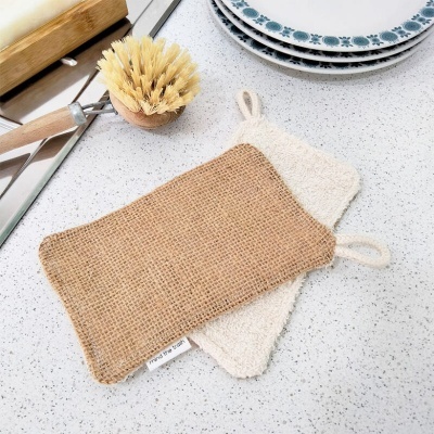 Dois panos de prato de ráfia e tecido felpudo sobre bancada branca com pratos e utensílios de cozinha