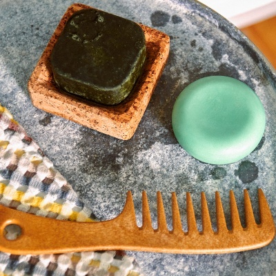 Sabonete quadrado escuro em base de cortiça, barra redonda verde e pente de madeira sobre superfície cinza