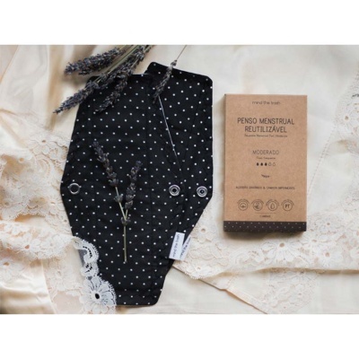 Penso menstrual reutilizável preto com pontos brancos ao lado de embalagem de papel castanho com texto em português