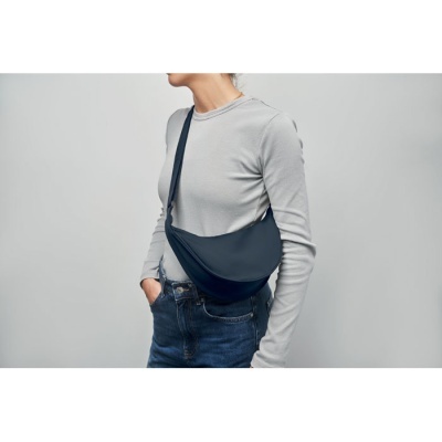 Pessoa com mala azul escura transversal, camisa cinzenta e jeans azuis sobre fundo cinzento.