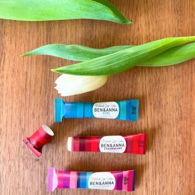 Três tubos de cosméticos Ben & Anna e carretel vermelho junto a tulipas brancas numa mesa de madeira