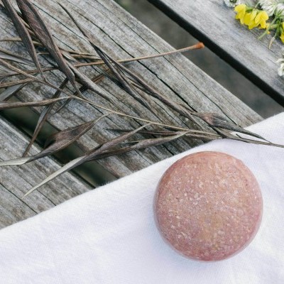 Barra redonda de xampu sólido rosa sobre pano branco e madeira com ramos secos e flores