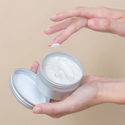 Frasco de creme branco com tampa prateada aberto em mãos femininas sobre fundo bege