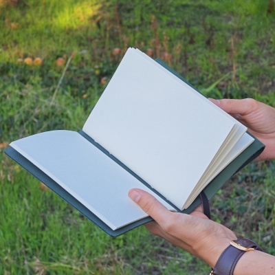 Caderno verde aberto com folhas em branco segurado por mãos ao ar livre