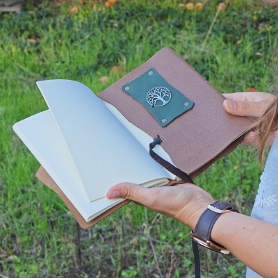 Caderno de capa castanha com placa verde e símbolo de árvore, páginas em branco, em mãos, fundo relva verde.