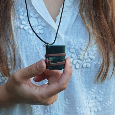 Colar com livro de couro verde em miniatura sendo segurado contra camisa branca bordada