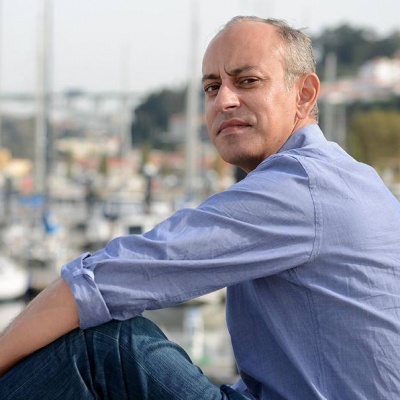 Homem com camisa azul clara e calças de ganga sentado perto de barcos num cais