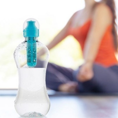 Garrafa de água com infusor azul e tampa azul, pessoa desfocada ao fundo sentada no chão