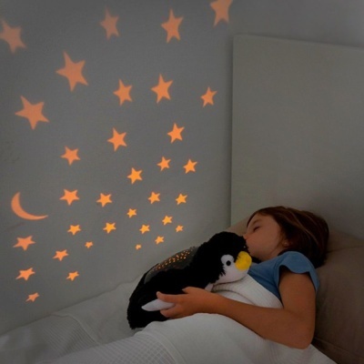 Criança de pijama azul abraçando peluche de pinguim na cama com estrelas laranja projetadas na parede