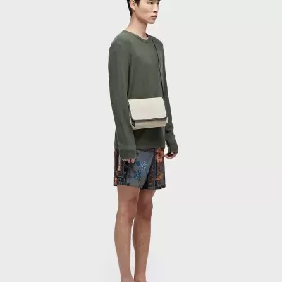 Homem com camisola verde, calções coloridos, chinelos pretos e mala bege a tiracolo