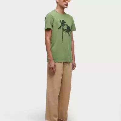 Homem com t-shirt verde com palmeiras, calças beje largas e sandálias pretas