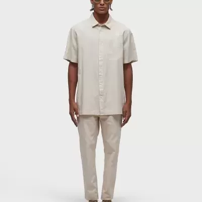 Homem de pé com camisa e calças bege, óculos de sol amarelos e sapatos castanho claro.