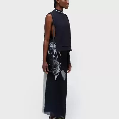 Modelo com top preto sem mangas e saia longa preta com flor cinzenta, calça tênis com detalhes verdes