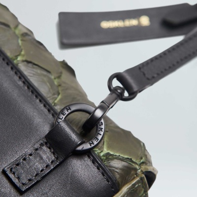 Saco verde com textura de pele de réptil e detalhes em pele preta, com argola metálica preta e etiqueta preta no fundo.