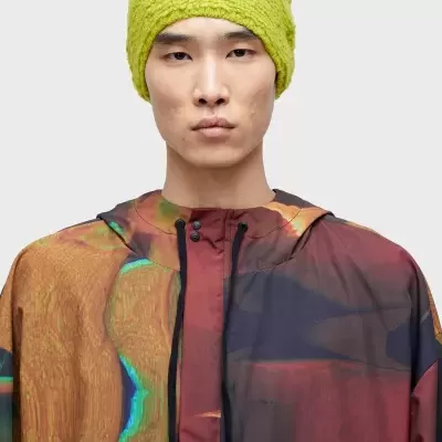 Homem com capa de chuva colorida e gorro verde limão num fundo cinza claro