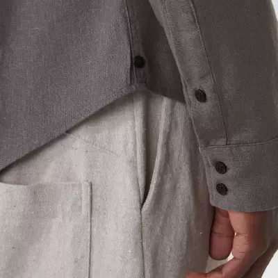 Camisa cinzenta com botões pretos e calças claras com bolso visível.