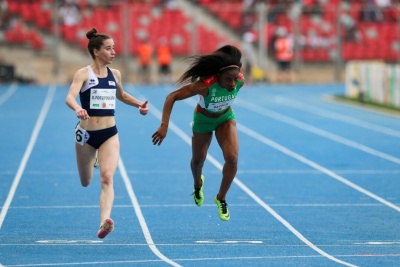 Duas atletas a correr numa pista de atletismo azul, vestem roupas desportivas, uma com o número 6 e a outra com roupa verde e sapatilhas amarelas.