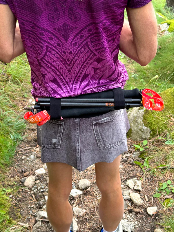 Pessoa com mochila preta e bastões de caminhada presa nas costas, em trilha com vegetação.