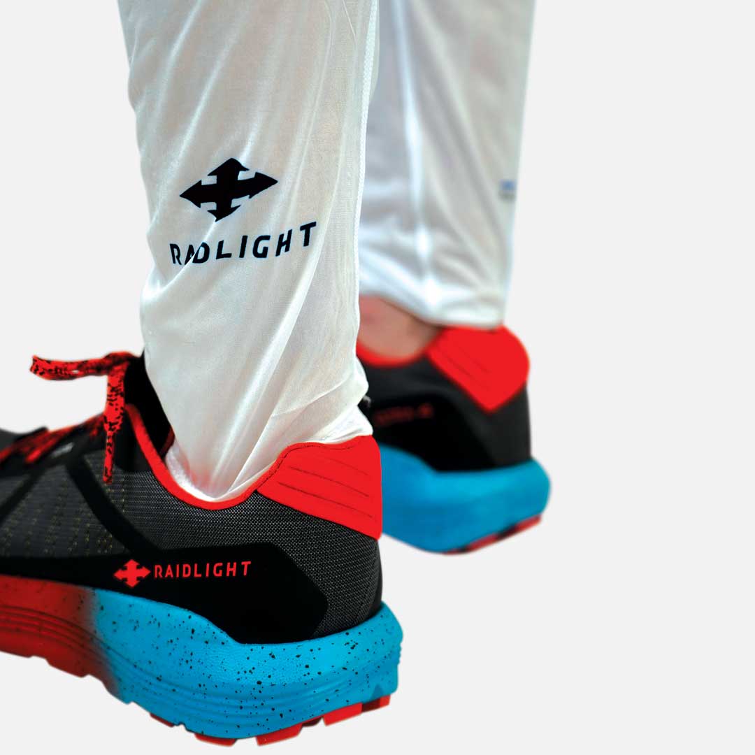 Sapatos desportivos Raidlight com base azul, detalhes vermelhos, corpo negro com cinzento e leggings brancas com logo Raidlight