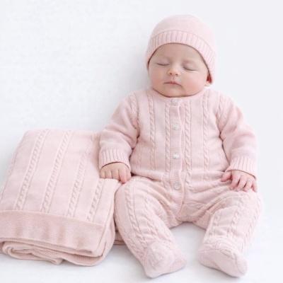Bebé com fato e gorro de malha rosa claro sentado junto a manta