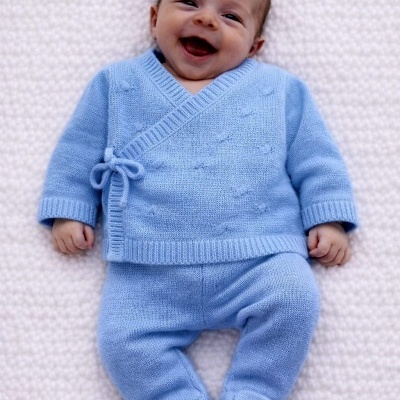 Bebé a sorrir vestido com conjunto de malha azul clara composto por camisola de laço e calças com pés.