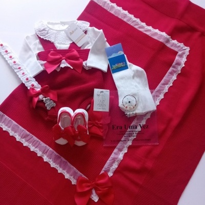Conjunto de roupa para bebé vermelho com laços e renda branca sobre manta vermelha.