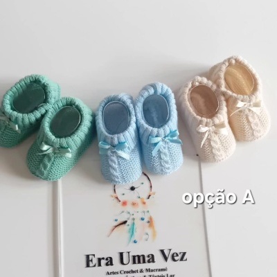 Três pares de sapatinhos de bebé em malha verde, azul claro e bege com laços, sobre placa com texto