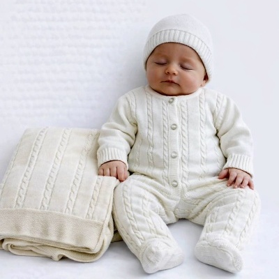 Bebé com fato de malha branco e gorro, junto a manta creme dobrada.